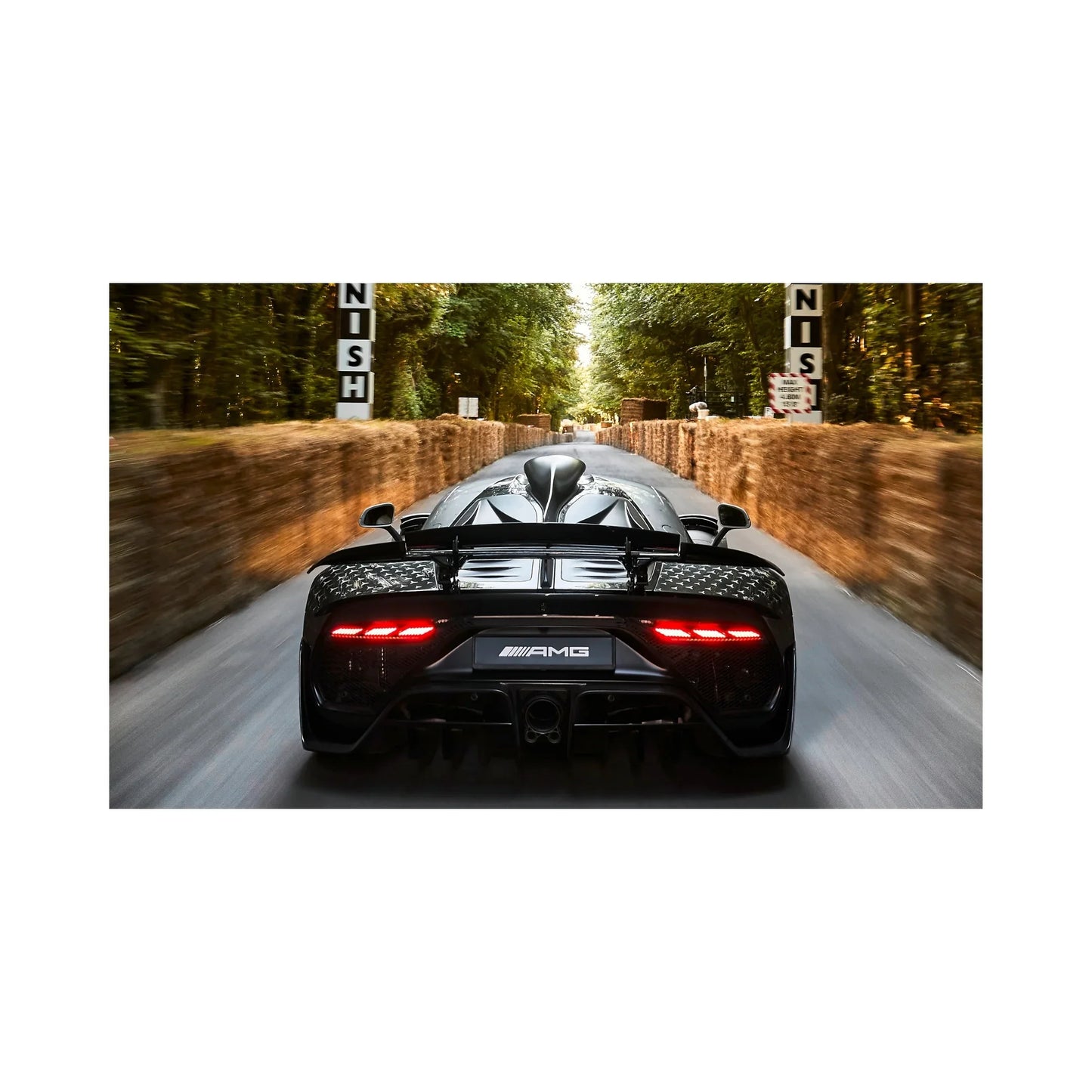 Rear view of a black Mercedes-AMG hypercar speeding on a tree-lined race track with hay bale barriers