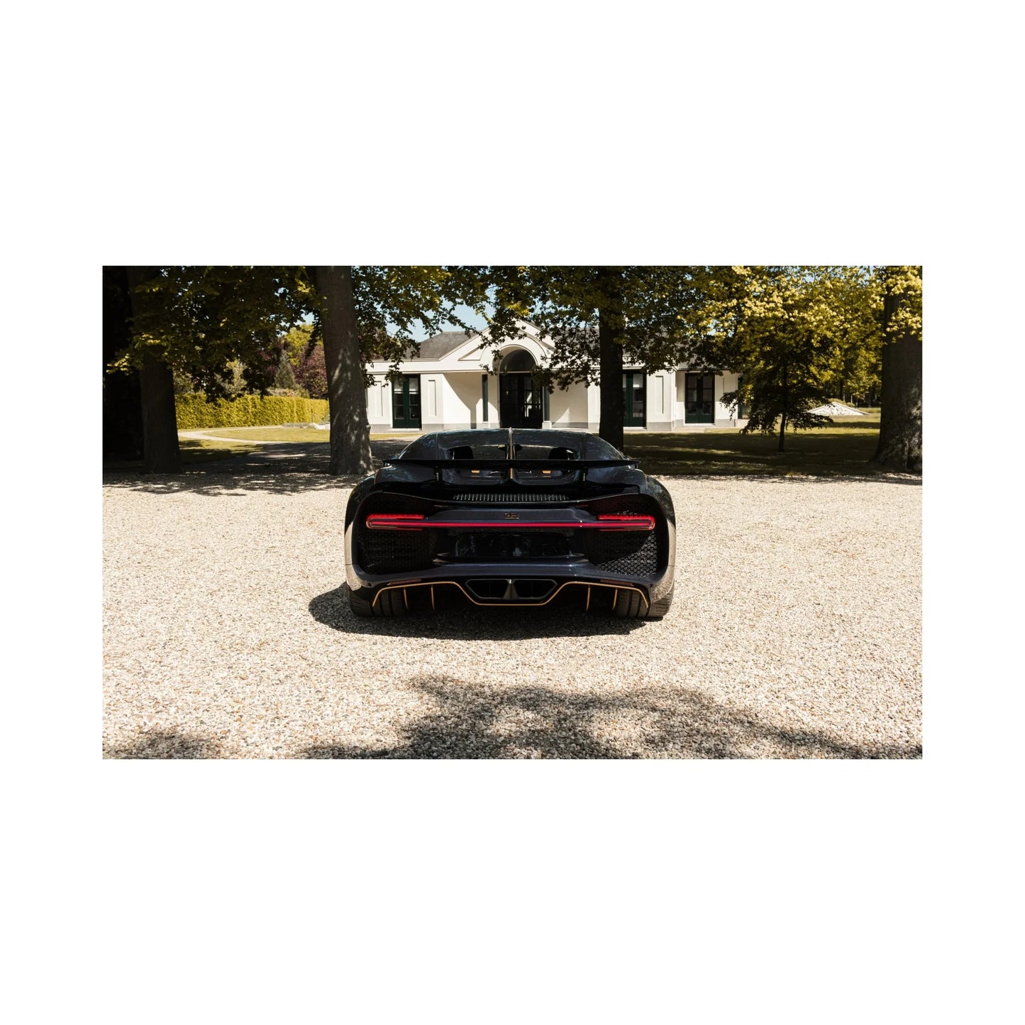 Rear view of a black luxury sports car on gravel driveway with a large white house and trees in background