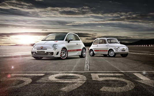Two white Fiat Abarth 595 cars, classic and modern, on an open road at sunset with cloudy sky
