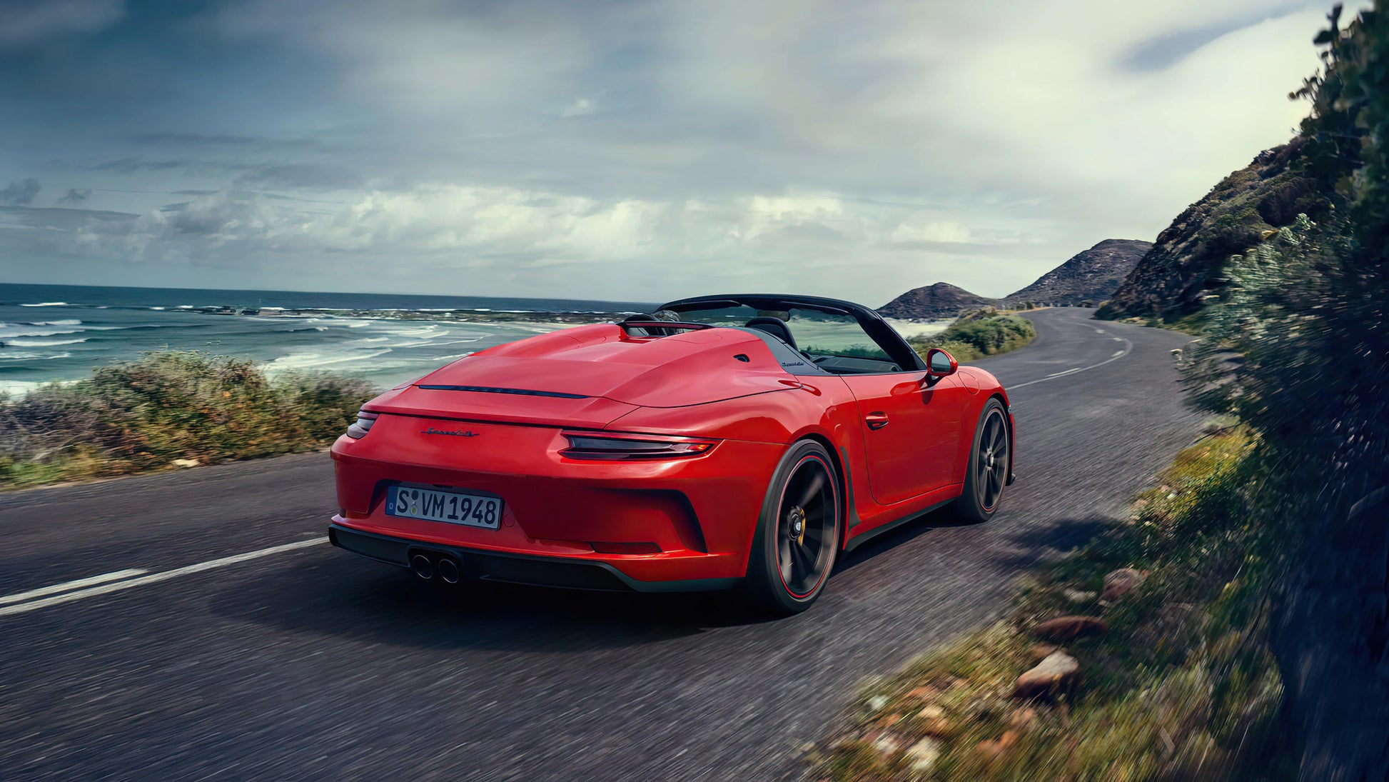 Red Porsche Speedster convertible driving on coastal road with ocean and hills