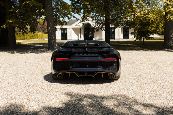 Rear view of black Bugatti Chiron parked on gravel in front of mansion, shaded by trees