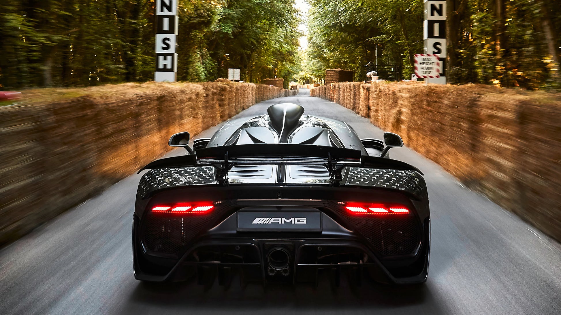 Rear view of black 2022 Mercedes AMG One hypercar speeding on forest road with hay bale barriers