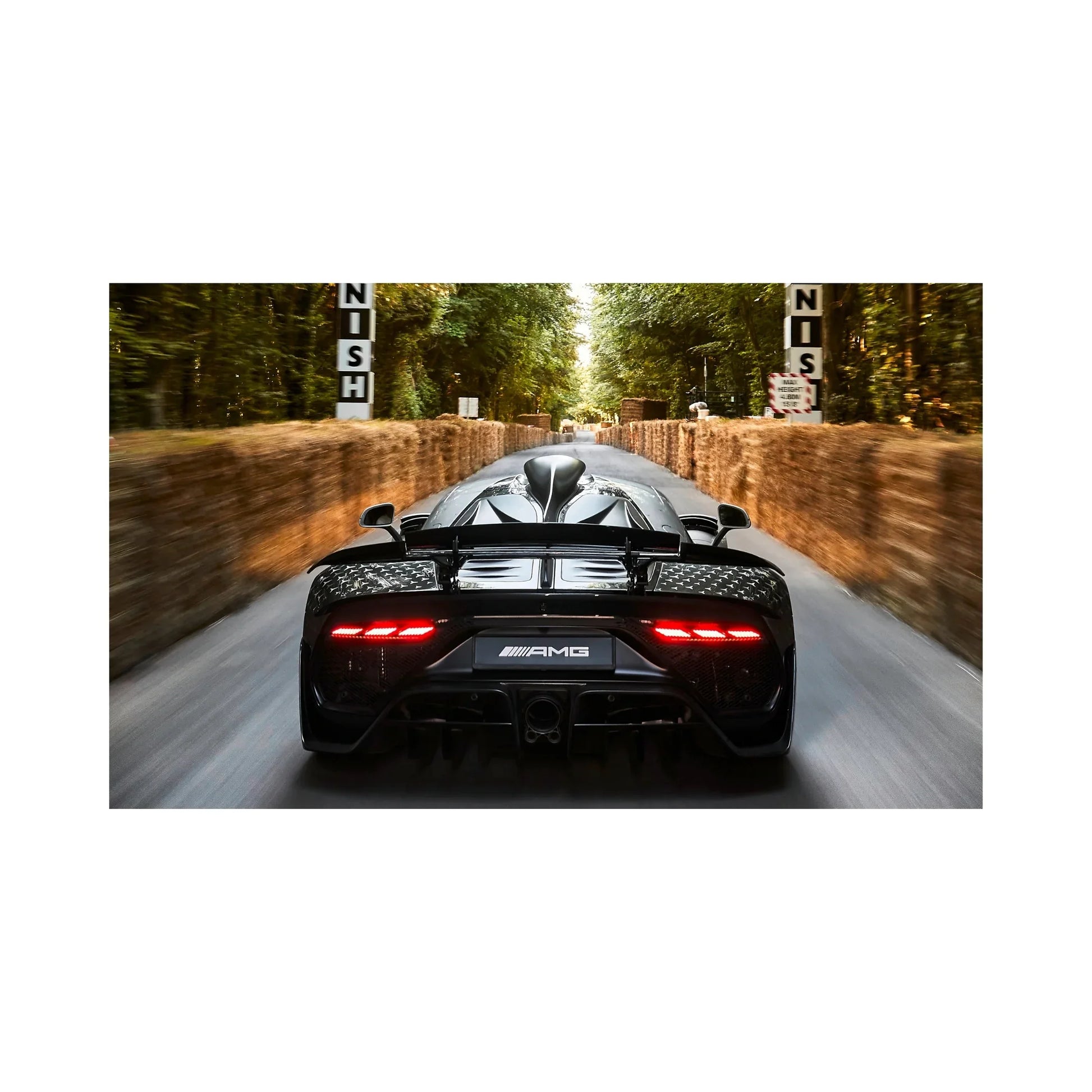 Rear view of a black Mercedes-AMG hypercar speeding on a tree-lined race track with hay bale barriers